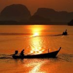 A man rows a longtail boat at sunset, with mountains framing the background.