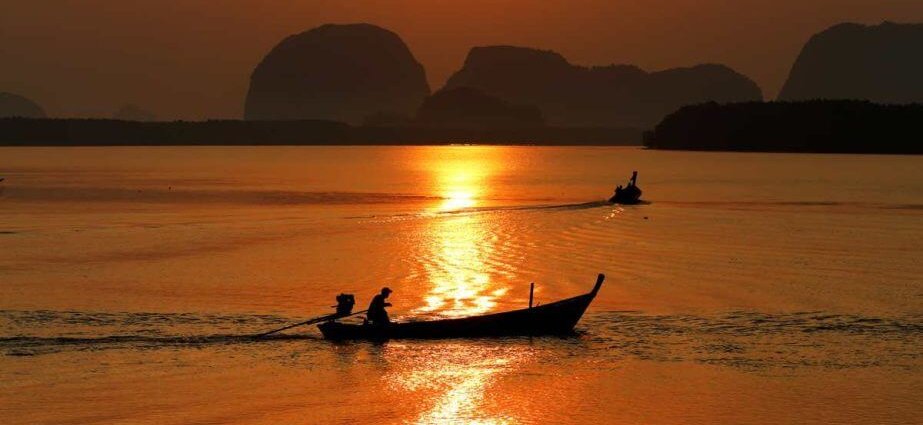 A man rows a longtail boat at sunset, with mountains framing the background.
