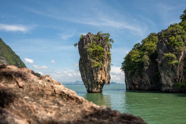 A tall limestone stack covered in greenery standing in the calm blue water of Phang Nga Bay, viewed from a rocky vantage point during a Simba Sea Trips Phang Nga Bay & Beyond tour.