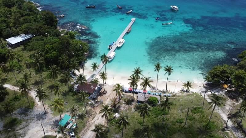 Aerial view of a tropical bay with turquoise water, a small pier, boats and palm‑fringed white‑sand beach.